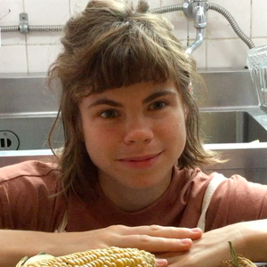 A portrait photo of me, Laurel Schwulst — I’m a white woman in my early 30s with medium length brown hair, and in this picture I’m wearing an apron and smiling in front of corn because I’m about to cook “summer corn soup” for my friends in Osaka Japan as part of my art residency at “pe hu,” a creative community space there. This photo was taken by Ayami Konishi, one of its members.